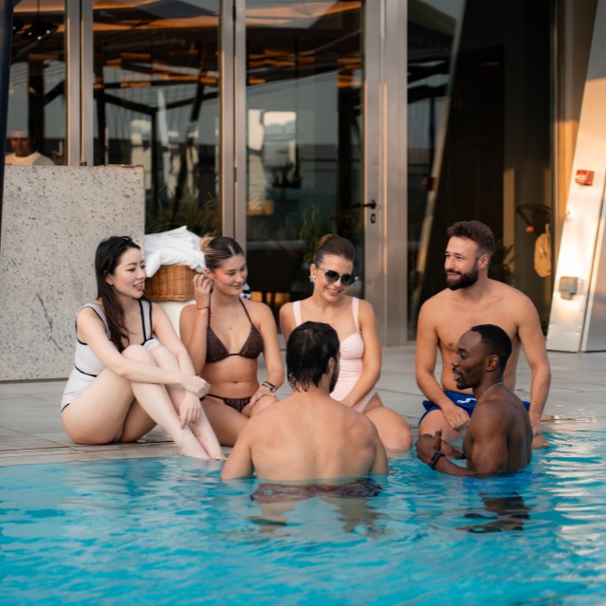 A group of six friends, four sitting at the pool's edge and two in the water, enjoy a relaxed conversation. They are smiling, creating a joyful, summer vibe.