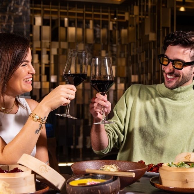 A woman and a man clink wine glasses together, surrounded by an elegant dining setup with various dishes on the table.