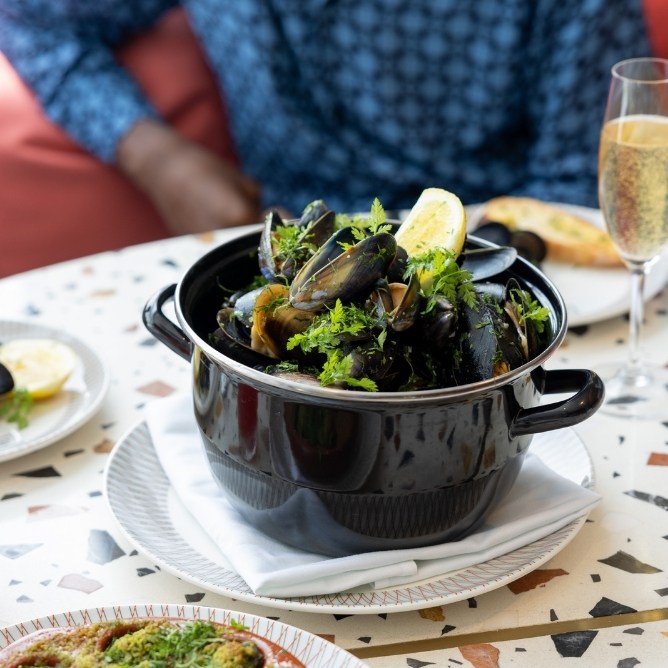 A black pot filled with mussels, garnished with lemon and herbs, sits on a table alongside a plate and a glass of bubbly drink.
