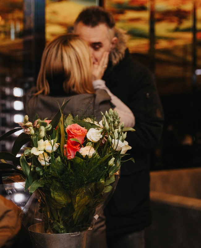 A bouquet with vibrant pink, white, and yellow flowers is in focus, while a couple shares an intimate moment in the blurred background at dusk.