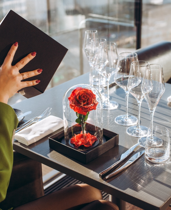 A preserved red rose encased in glass sits on a restaurant table set with wine glasses and cutlery. A person holds a menu. The scene is elegant and romantic.