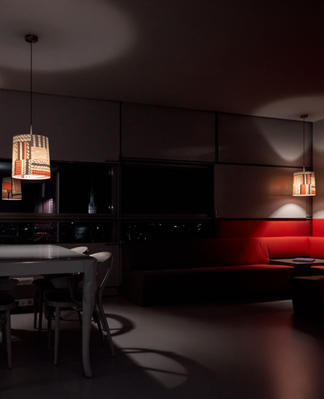 A dimly lit modern room features patterned pendant lights, a white dining table with chairs, and a red corner sofa. City lights are visible through the window. The mood is cozy and serene.