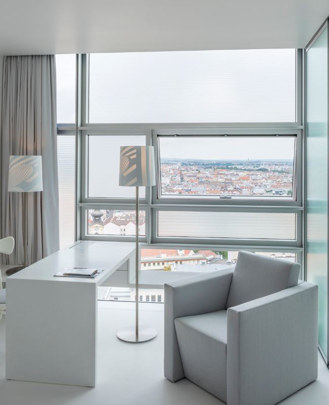 Modern room with a large window view of a sprawling cityscape. A sleek white desk, floor lamp, and cushioned armchair create a minimalist and serene atmosphere.