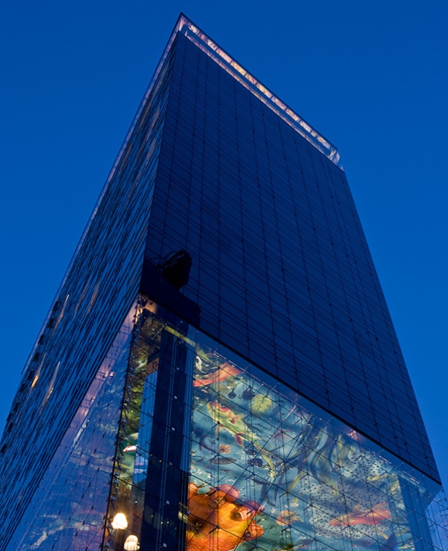 A modern glass skyscraper with vibrant, colorful art displayed on its lower facade, set against a deep blue twilight sky, conveys a dynamic, urban atmosphere.