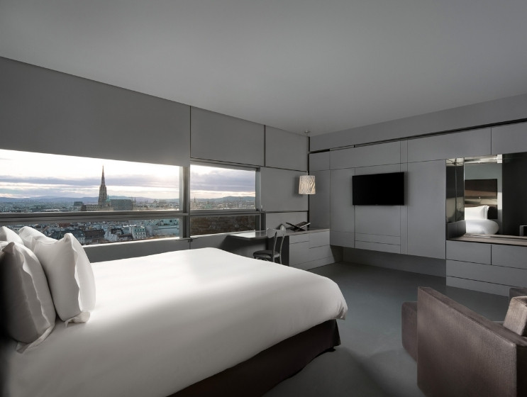 Modern hotel room with a large bed facing floor-to-ceiling windows overlooking a city skyline and cathedral. Sleek gray decor, cozy ambiance.