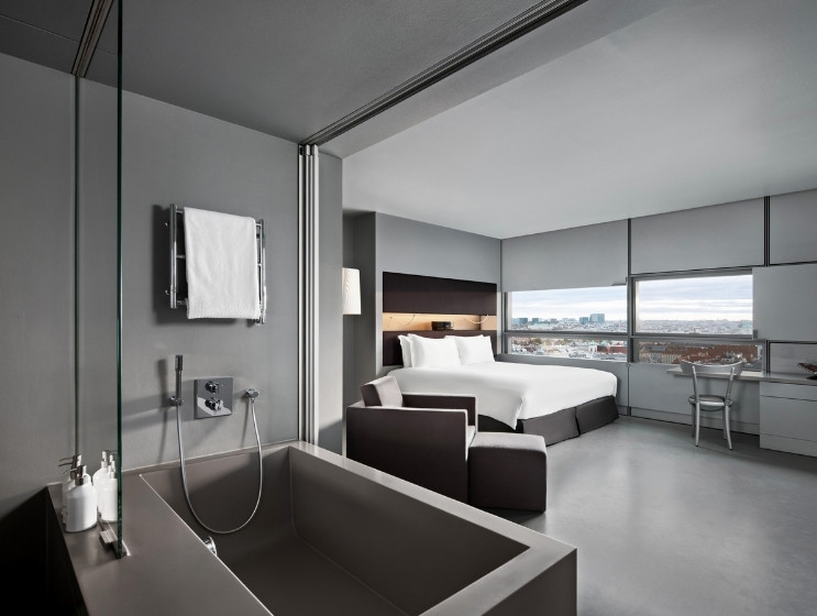Modern hotel room with minimalist design. Features a bathtub, sleek bed, large windows with city view, and neutral gray tones creating a calm, sophisticated atmosphere.