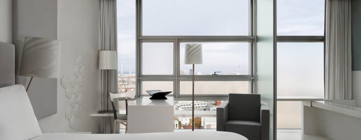 Modern, minimalist hotel room with large windows showcasing a city view. Features sleek white furniture, gray armchair, and soft lighting, creating a calm ambiance.