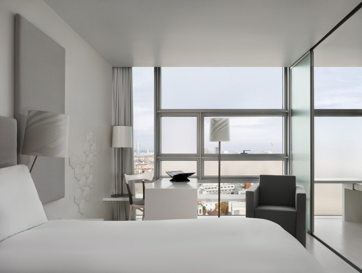A modern hotel room with minimalist design featuring a large bed, glass wall, sleek furniture, and a window offering a view of the city skyline.