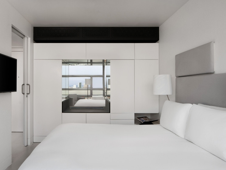Modern hotel room with a white bed, wall-mounted TV, and large windows reflecting a cityscape. The ambiance is clean and minimalist.