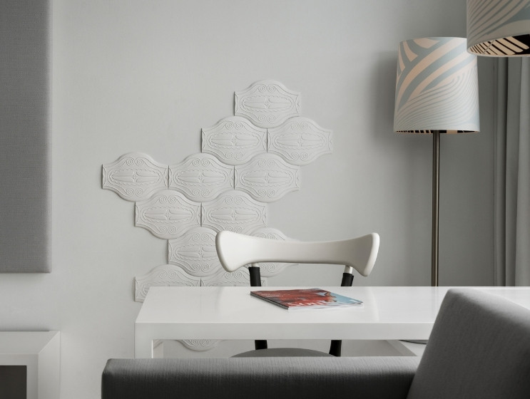 Modern, minimalist room with white wall tiles forming a pattern, white desk, sleek white chair, and a floor lamp with a geometric shade. Elegant and serene ambiance.