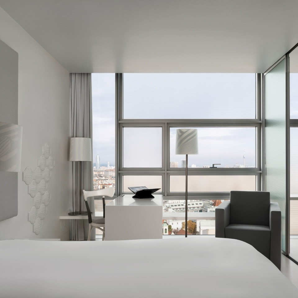 Modern, minimalist hotel room with large windows showcasing a city view. Features sleek white furniture, gray armchair, and soft lighting, creating a calm ambiance.