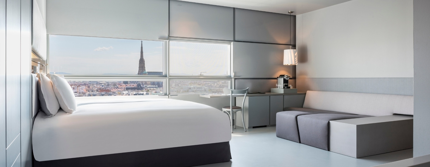 Modern hotel room with large bed, gray sofa, desk, and chair. A window offers a city view with a prominent tower. Neutral tones create a calm atmosphere.