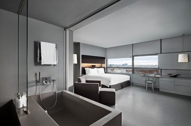 http://Modern%20hotel%20room%20with%20minimalist%20design.%20Features%20a%20bathtub,%20sleek%20bed,%20large%20windows%20with%20city%20view,%20and%20neutral%20gray%20tones%20creating%20a%20calm,%20sophisticated%20atmosphere.