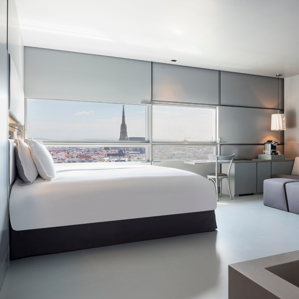 Modern hotel room with large bed, gray sofa, desk, and chair. A window offers a city view with a prominent tower. Neutral tones create a calm atmosphere.