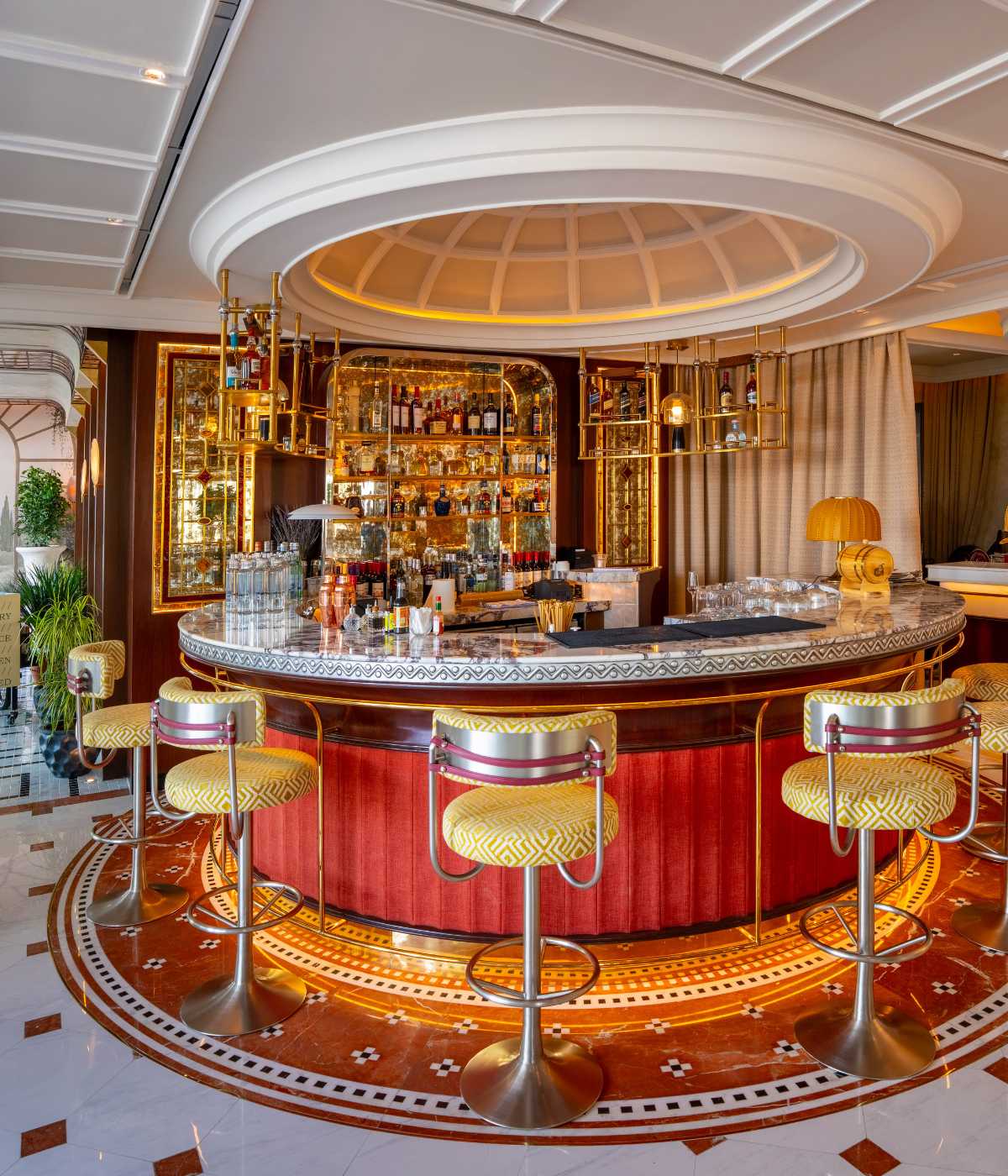 Luxurious bar with a circular counter, ornate design, and red cushioned stools. Bottles and glassware display elegance under soft, warm lighting.