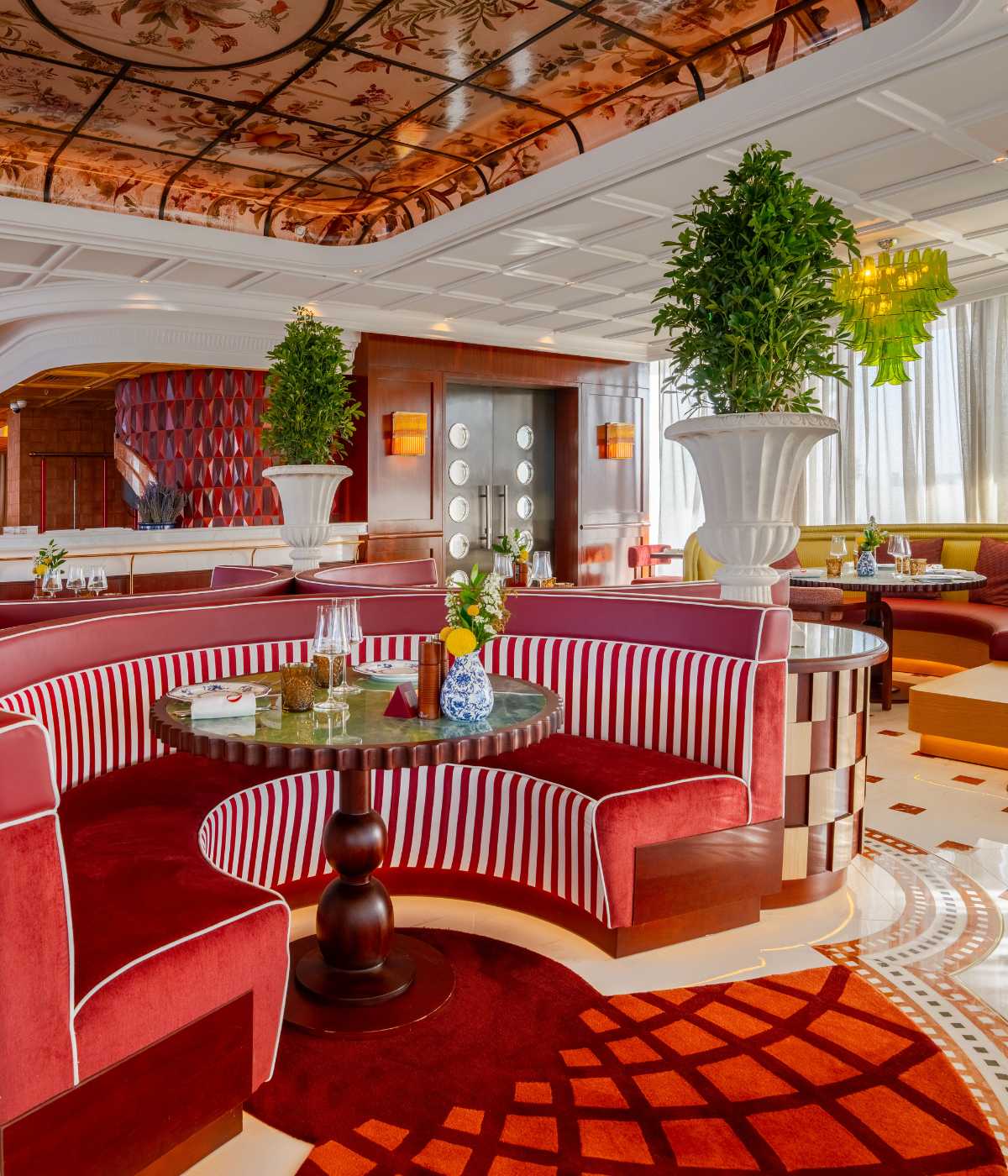 Luxurious restaurant interior with plush red striped booths, elegant marble tables, floral ceiling, and large potted plants, creating a vibrant, upscale ambiance.