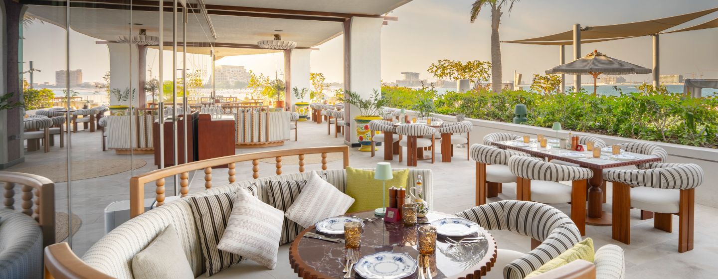 Rooftop restaurant with elegant curved seating, set for dinner. Tables have striped cushions and glasses, surrounded by greenery at sunset.