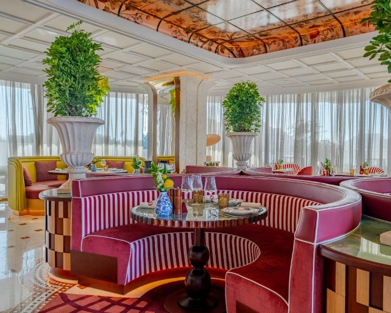 http://Luxurious%20restaurant%20interior%20with%20round%20red%20velvet%20booths,%20large%20potted%20plants,%20and%20a%20warm,%20elegant%20ambiance.%20Sunlight%20filters%20through%20sheer%20curtains.