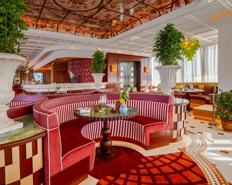 http://Luxurious%20restaurant%20with%20elegant%20red%20banquette%20seating,%20green%20plants%20in%20large%20vases,%20artful%20ceiling,%20and%20sunlight%20streaming%20through%20curtains.