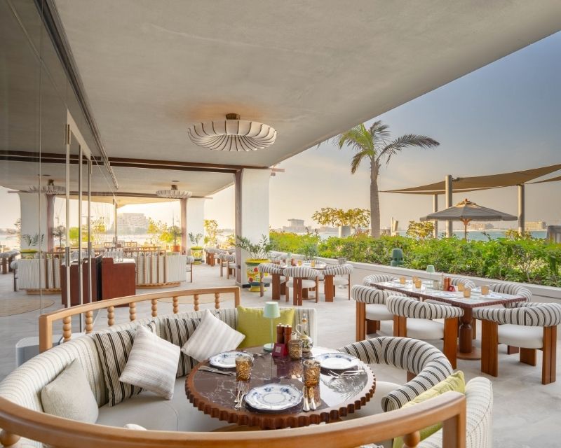 http://Outdoor%20restaurant%20terrace%20with%20modern%20round%20seating%20and%20wooden%20tables.%20Overlooks%20lush%20greenery%20and%20distant%20cityscape%20under%20a%20clear,%20sunny%20sky.%20Cozy%20and%20inviting%20atmosphere.