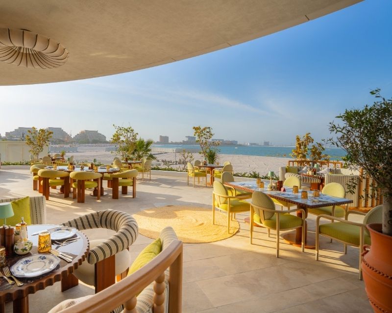 http://Beachfront%20restaurant%20patio%20with%20stylish%20green%20and%20yellow%20decor,%20overlooking%20an%20ocean%20view.%20Tables%20set%20for%20dining,%20sunlit,%20relaxing%20atmosphere.