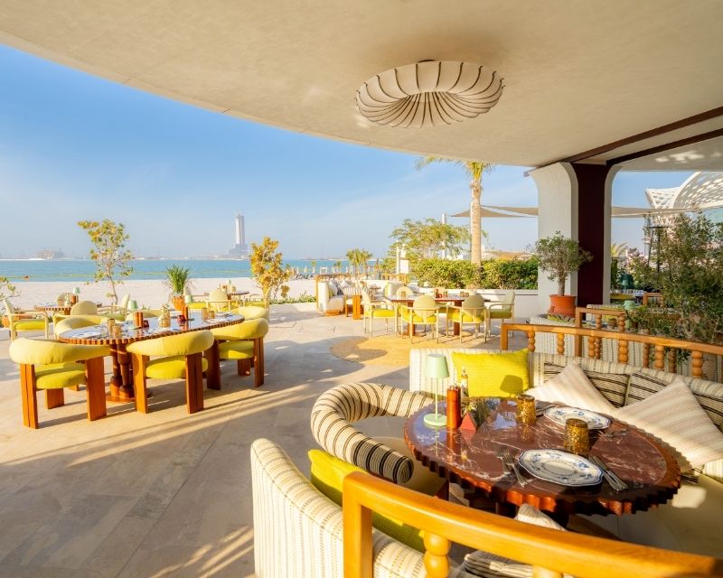http://Outdoor%20beachfront%20patio%20with%20yellow%20and%20green%20cushioned%20seating%20and%20round%20tables%20set%20for%20dining.%20Ocean%20view%20and%20clear%20blue%20sky%20create%20a%20relaxing%20atmosphere.