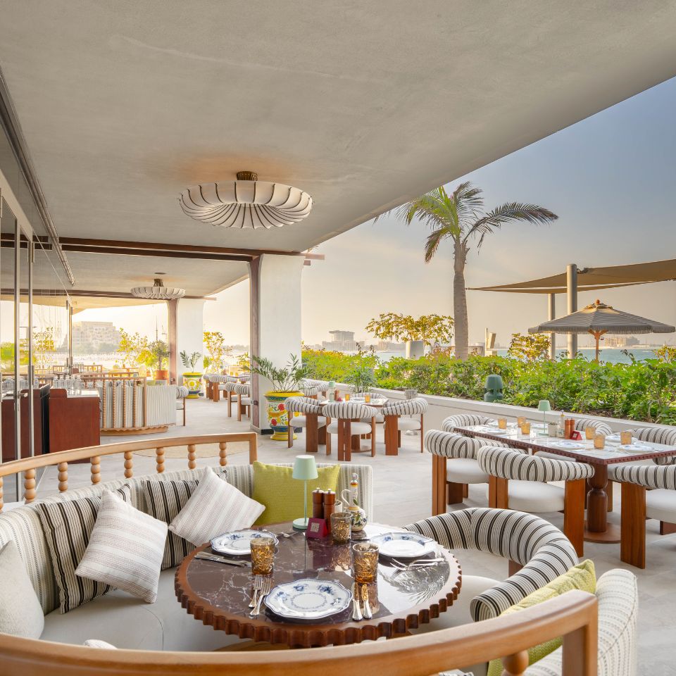 Rooftop restaurant with elegant curved seating, set for dinner. Tables have striped cushions and glasses, surrounded by greenery at sunset.