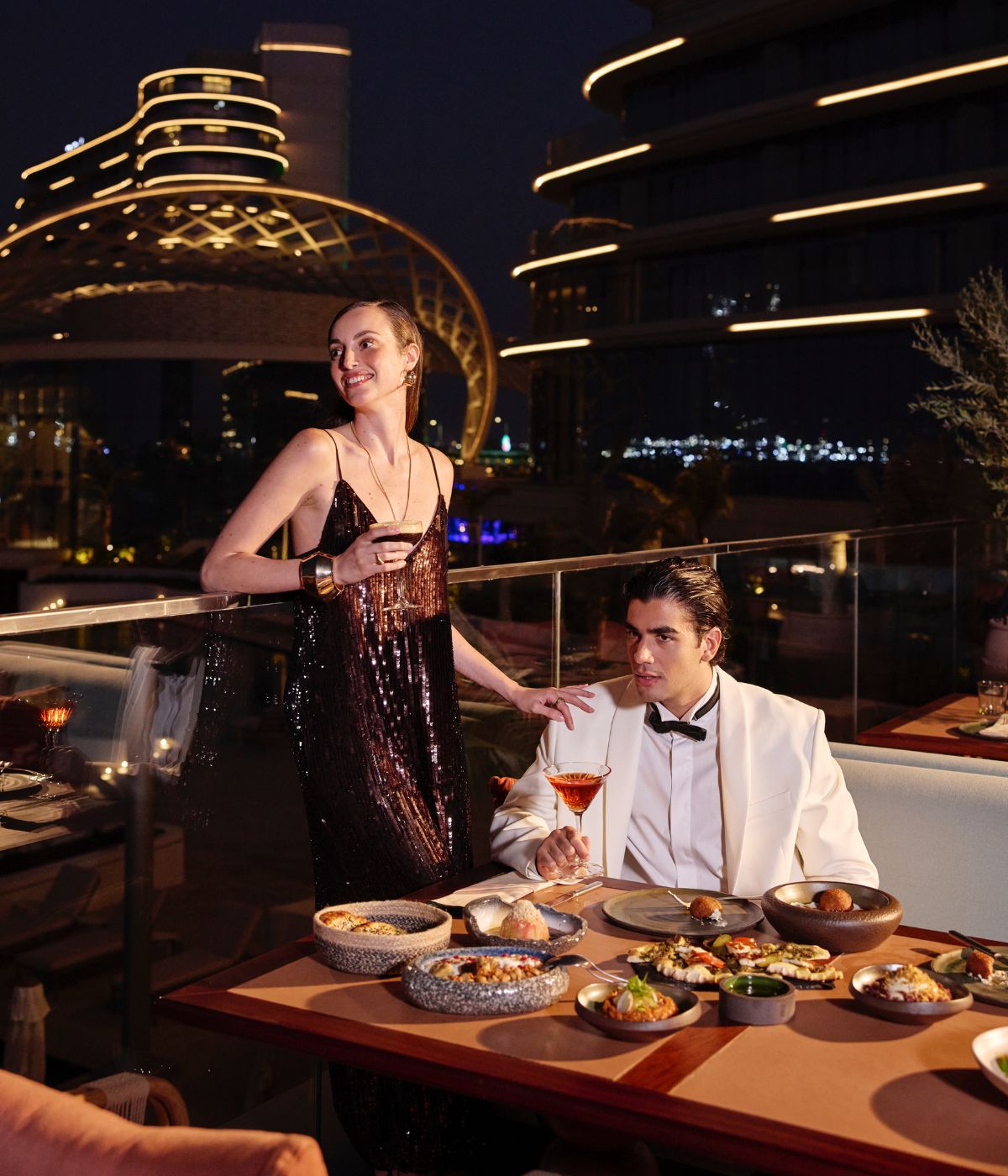 A glamorous couple at a rooftop restaurant at night. A woman in a sparkling dress holds a drink, while a man in a tuxedo sits relaxed, holding a cocktail. City lights and modern architecture glow in the background, creating a luxurious ambiance.