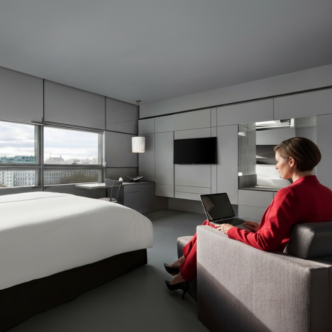 A modern hotel room featuring minimalistic design, with a large window, a bed, and a person working on a laptop in a suit.