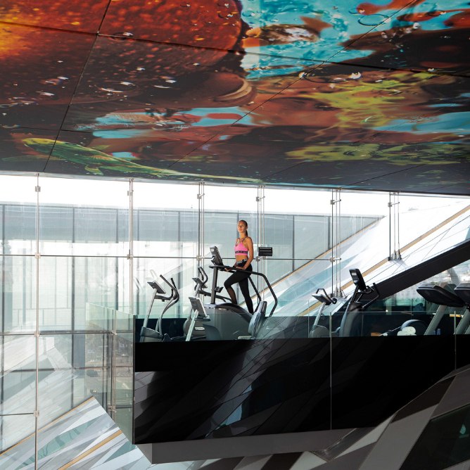 A woman exercises on an elliptical in a modern gym with a vibrant underwater-themed ceiling above her.