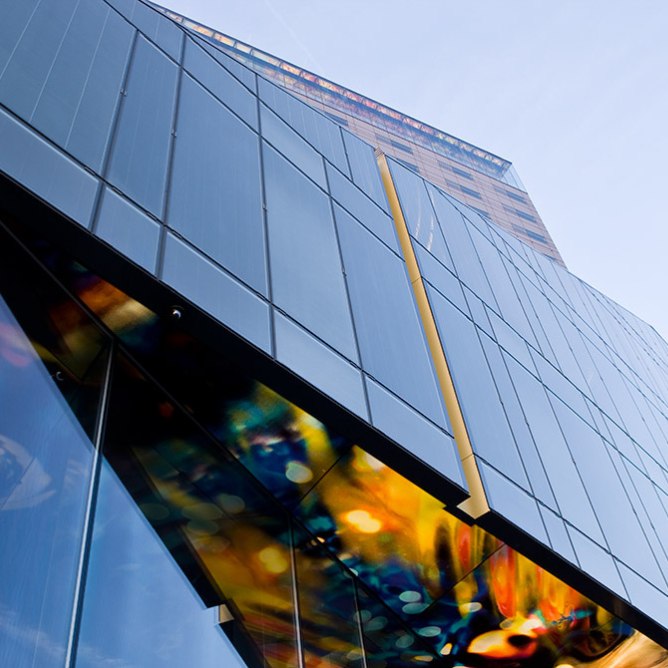 Skyscraper with reflective glass panels, a yellow vertical stripe, and a colorful abstract pattern at the base against a blue sky backdrop.