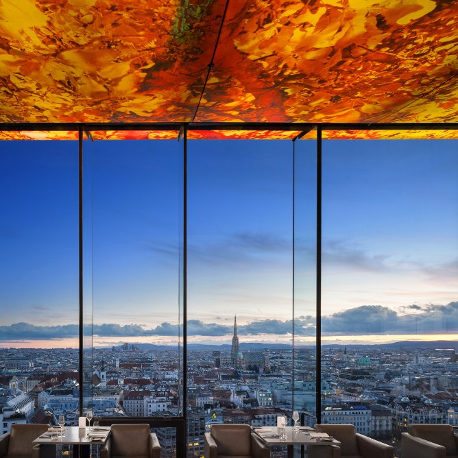 Interior view of a modern restaurant with a colorful ceiling, showcasing a panoramic sunset over Vienna’s skyline.