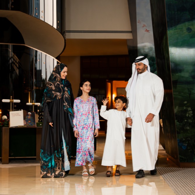 A family of four walking indoors; a woman in a patterned abaya and man in white traditional attire, with two children in colorful outfits, smiling warmly.