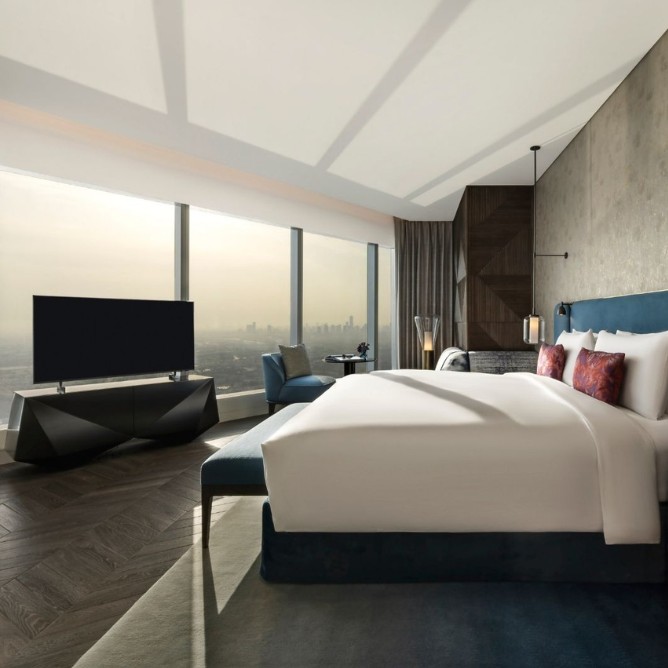 Modern hotel room with floor-to-ceiling windows offering a city skyline view. Features a king-sized bed, flat-screen TV, armchair, and elegant decor.