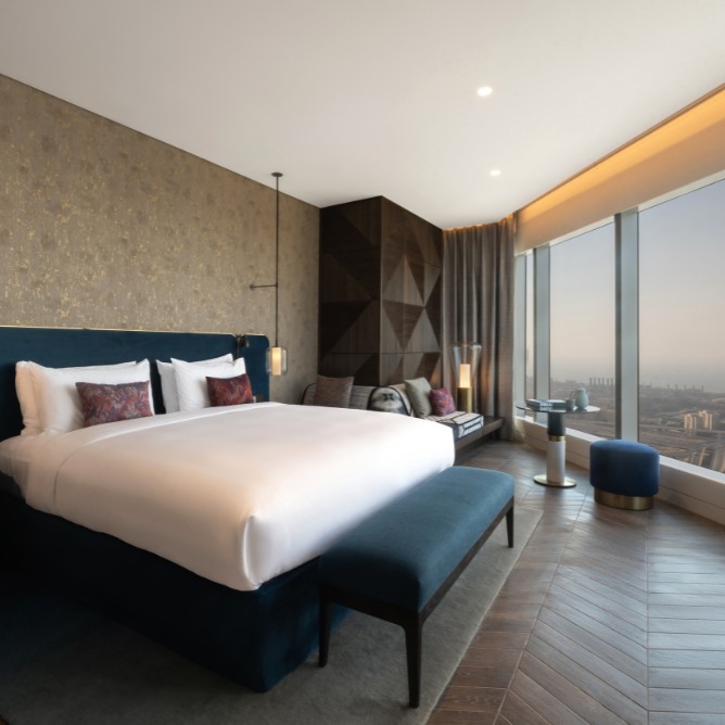 Modern hotel room with a plush king-size bed, blue bench, elegant gold-accented walls, floor-to-ceiling windows, city view, and cozy seating area.