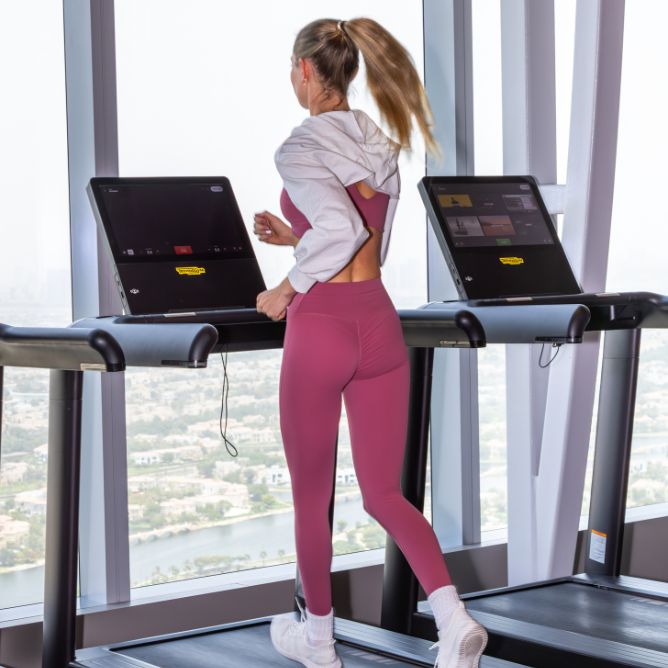 A woman in pink activewear jogs on a treadmill by a large window with a city view. The scene conveys energy and focus in a modern gym setting.