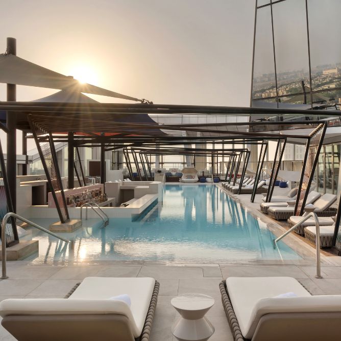 Rooftop pool area with sun loungers, a bar, and modern architecture, basking in golden sunlight against a city backdrop.