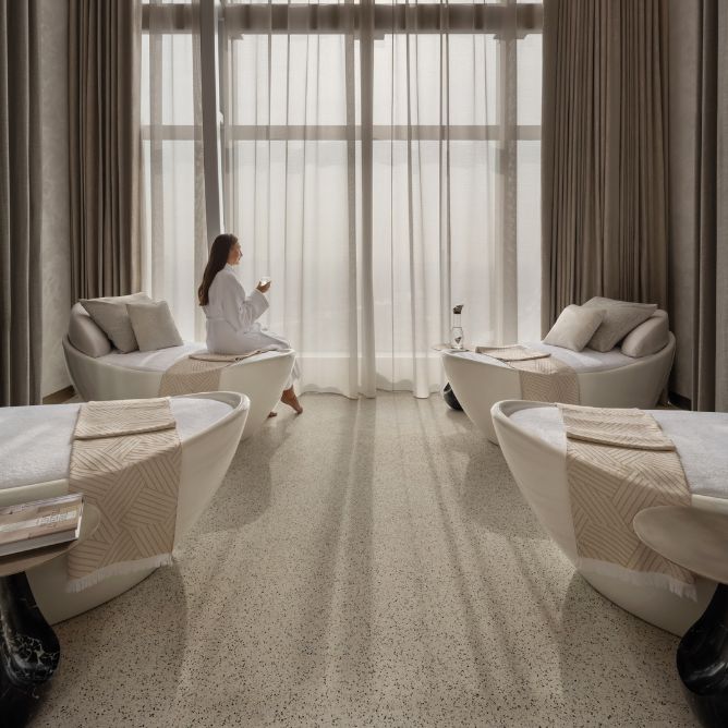 A serene spa room with a relaxed woman in a robe, gazing out through sheer curtains, surrounded by elegant treatment chairs.
