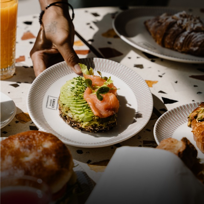 A hand holds a plate with avocado toast topped with smoked salmon and greens, surrounded by breakfast items and a glass of orange juice.