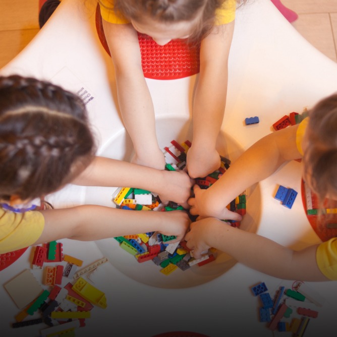Three children in yellow shirts eagerly reach into a bowl filled with colorful LEGO bricks, showcasing playfulness and creativity in a collaborative setting.