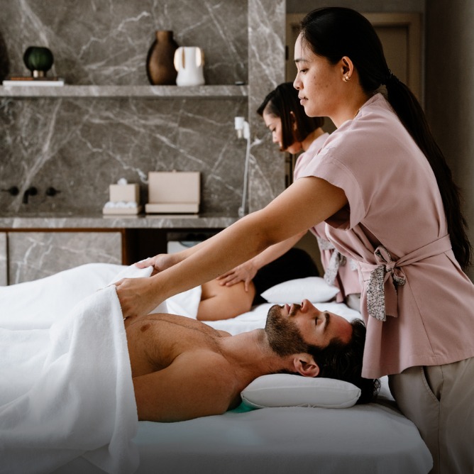 Two people receive relaxing massages on white beds. Masseuses in pink uniforms focus intently in a serene spa with stone wall decor.