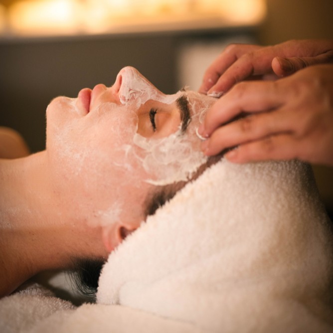 A woman with a towel wrapped around her head receives a relaxing facial massage, her face covered in a soothing white mask, in a serene spa setting.