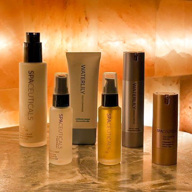 A collection of six skincare products on a marble surface, warmly lit from behind. The bottles and tubes feature the brands Spaceuticals and Waterlily.