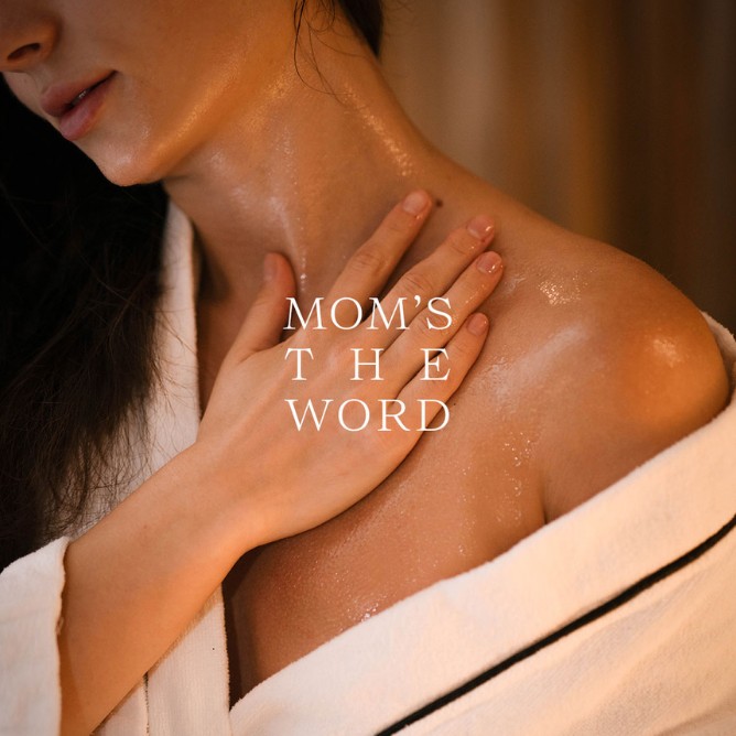 A woman in a bathrobe gently touches her collarbone, with moisturized skin glistening. The text "Mom's the Word" overlays the image, conveying warmth and care.