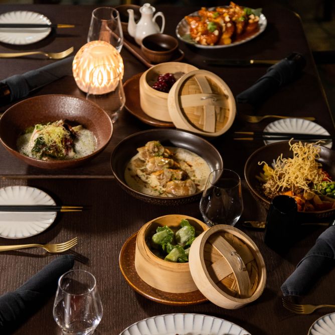 A candlelit dining table set with elegant dishes, including dumplings, green vegetables, and meat platters, creating a warm, inviting ambiance.