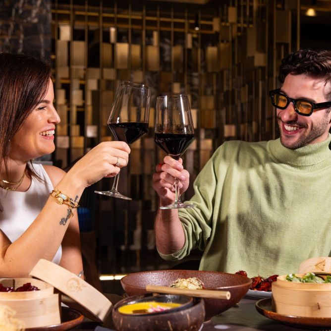 A man and woman toast with red wine glasses, smiling warmly at a restaurant. The table is set with food, capturing a joyful, relaxed ambiance.