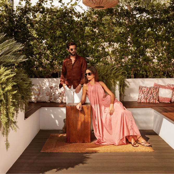 Two people in stylish outfits posing on a wooden deck with white seating and green plants in the background.