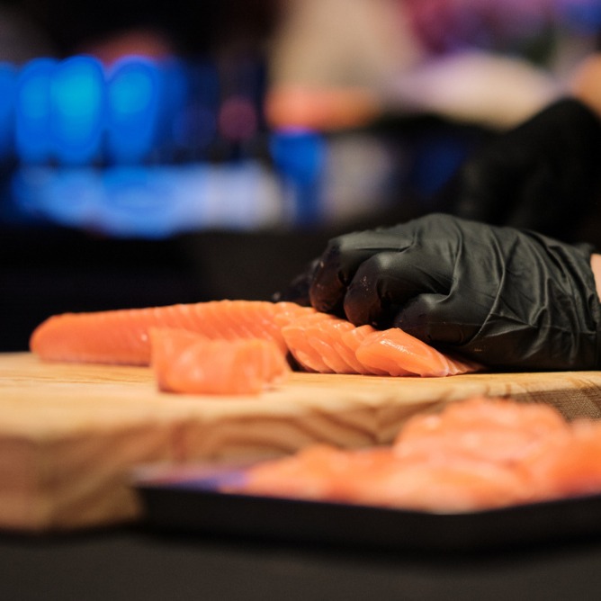 A person wearing black gloves slices fresh salmon on a wooden board. The background is blurred with blue lights, creating a calm and focused atmosphere.