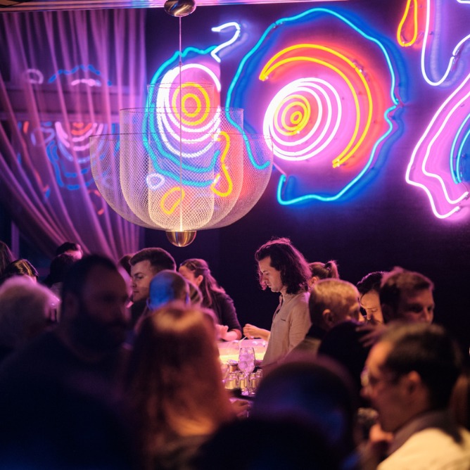Dimly lit bar with vibrant neon abstract wall art. Patrons converse under soft overhead lights, creating a lively, modern atmosphere.