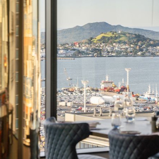 View from a restaurant, showing a harbor, ships, and distant hills under a clear sky. A table with blue chairs and set dining ware in the foreground.